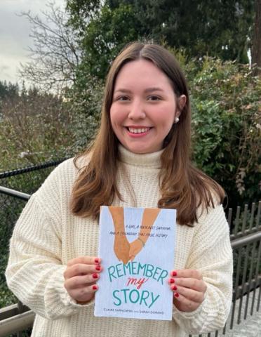 Claire Sarnowski has long brown hair in a white sweater while holding her copy of Remember My Story in her hands.