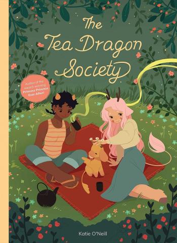 The book cover of The Tea Dragon Society. The background setting is a meadow with two characters sitting on a red blanket with a chamomile dragon in between them. One of the characters is holding a hot tea.  